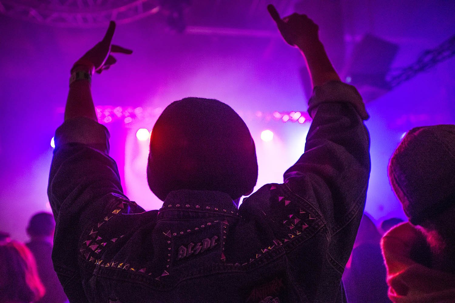 Back of a man at a concert. Hes wearing a beanie hat and a baggy denim jacket with studs. Photo with a purple hue